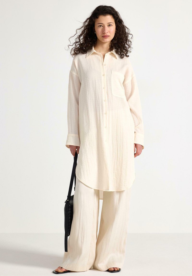 Camicia button-up lunga beige con tasca, abbinata a pantaloni beige a gamba larga. Tessuto leggero e testurizzato con un delicato motivo a righe.