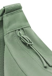 Angolo di una borsa in tela verde con un esterno testurizzato, dotata di una cerniera e un tirante, che mostra una finitura opaca e un design strutturato.