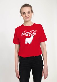 Rød bomulds T-shirt med et hvidt Coca-Cola isbjørn grafik og teksten "ENJOY" i kursiv skrifttype. Kortærmet og med rund halsudskæring.