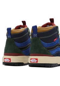 Un par de zapatillas altas Vans en azul, verde y beige con logo rojo, suelas negras y un interior forrado de felpa visto desde la parte de atrás.