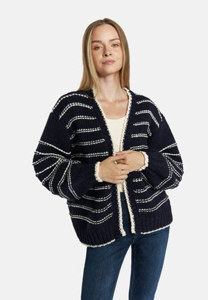 Jonge vrouw met lang blond haar draagt een marineblauwe grofgebreide cardigan met witte horizontale strepen en witte bies, gecombineerd met een blauwe spijkerbroek.