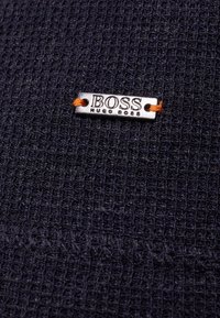 Navy textured fabric with a woven pattern, featuring a silver metal tag that reads "BOSS" attached with an orange thread.