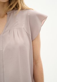 Blusa di un tenue malva con maniche corte a cappuccio, caratterizzata da pieghe sottili, scollatura a V e una texture liscia. Nessun motivo o hardware visibile.