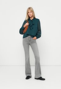 Τυρκουάζ μπλούζα από chiffon με φουσκωτές λεπτομέρειες, συνδυασμένη με ανοιχτό γκρίζα καμπάνα τζιν και μαύρες παπούτσια. Το μοντέλο κρατάει μια καφέ τσάντα χιαστί.