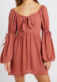 Růžovo-červené šaty s designem bez ramínek, detailem na zavazování u výstřihu, nařasenými rukávy a splývavou sukní vyrobenou z lehké, strukturované látky.