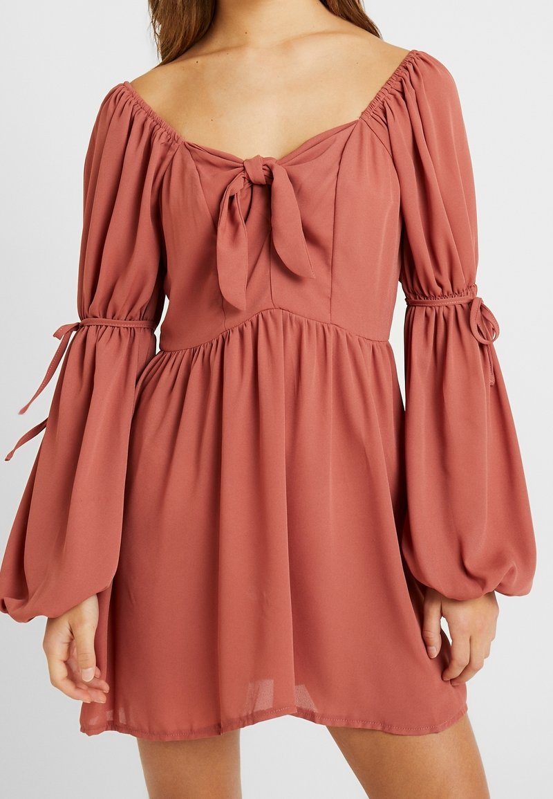 Růžovo-červené šaty s designem bez ramínek, detailem na zavazování u výstřihu, nařasenými rukávy a splývavou sukní vyrobenou z lehké, strukturované látky.
