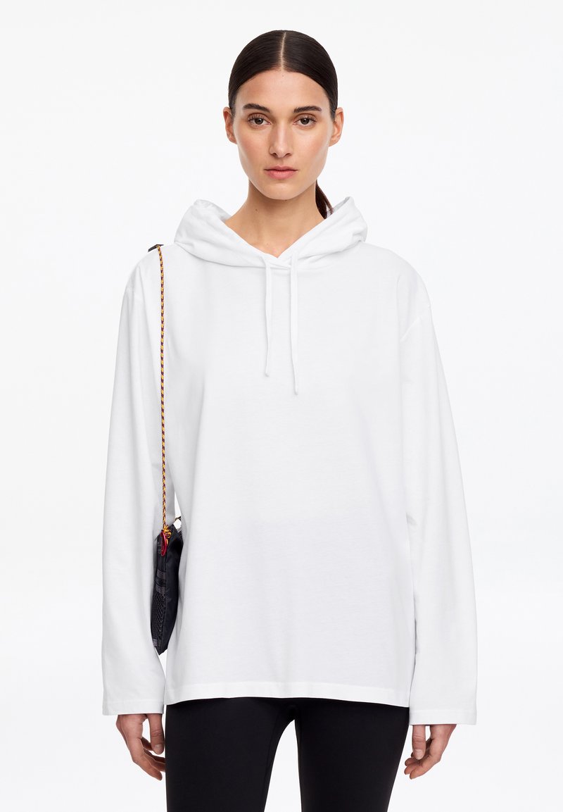 Jeune femme aux cheveux tirés en arrière porte un sweat à capuche blanc uni et un pantalon noir, portant un petit sac à bandoulière noir avec une sangle multicolore.
