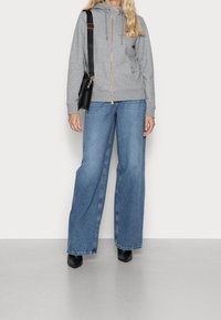 Un hoodie gris zippé avec une capuche, associé à un pantalon en denim bleu à jambes larges. Des bottines noires et un sac à épaule complètent l'ensemble.