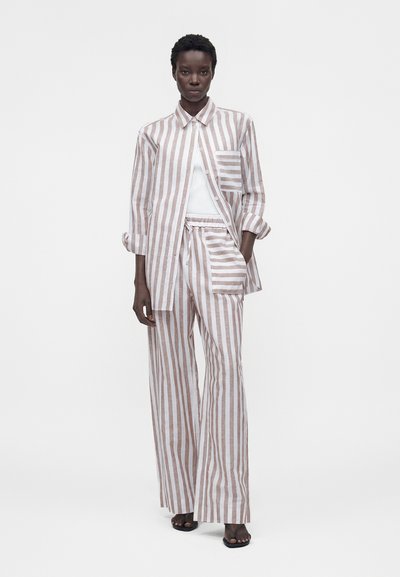 Ψηλό άτομο που φοράει μπεζ και λευκό κάθετο ριγέ πουκάμισο με κουμπιά και ασορτί φαρδιά παντελόνια, ποζάροντας με το ένα χέρι στην τσέπη.