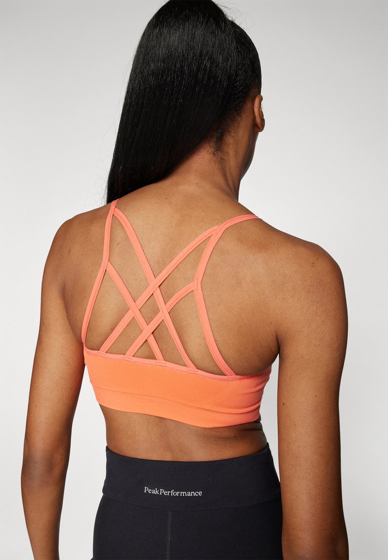 Oranger Sport-BH mit einem Kreuz-Design hinten und glatter Textur. Getragen mit schwarzen High-Waist-Leggings.