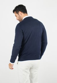Pull bleu marine à manches longues, avec des poignets et un ourlet côtelés, texture lisse et design classique à col rond. Associé à un pantalon de couleur claire.