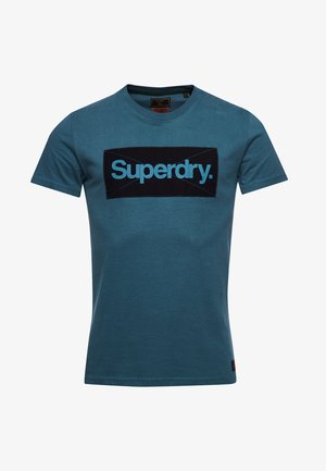 Blaues T-Shirt mit kurzen Ärmeln und Rundhalsausschnitt mit einem schwarzen rechteckigen Aufnäher und türkisfarbenem "Superdry."-Schriftzug zentriert auf der Brust.