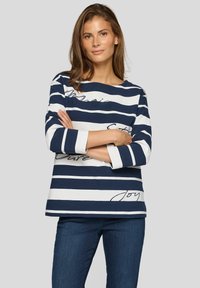 Frau mit braunen Haaren steht mit verschränkten Armen und trägt ein langärmliges, navyblau-weiß gestreiftes Hemd und Jeans vor einem grauen Hintergrund.