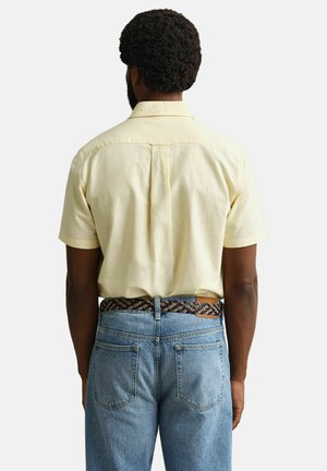 Mann mit lockigem Haar, der ein blassgelbes Kurzarmhemd trägt, das in eine blaue Jeans mit einem gemusterten braunen Gürtel gesteckt ist, von hinten betrachtet.