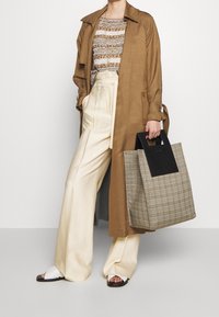 Brauner, langer Trenchcoat, gestreiftes Strickoberteil, cremefarbene weite Hose, schwarz-beige kariertete Tasche mit einem einfarbigen schwarzen Griff, weiße Rutschsandalen.
