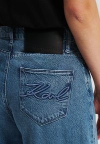 Blaue Jeans aus Denim mit einer diamantförmigen Tasche, die mit "Karl" in dunkelblau gestickt ist, sowie einem schwarzen Lederetikett am Bund.