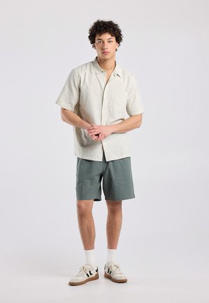 Joven con cabello rizado viste una camisa beige claro de manga corta, pantalones cortos verdes, calcetines blancos y zapatillas blancas con detalles negros.
