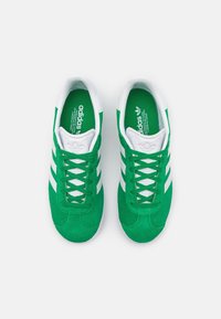 Baskets en daim vert avec des lacets blancs, trois rayures blanches et une pointe texturée. L'intérieur possède une semelle intérieure verte avec logo.