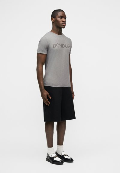 Άνδρας που στέκεται κοιτώντας μπροστά, φορά γκρι μπλουζάκι με την επιγραφή "DONDUP", μαύρα σορτς μέχρι το γόνατο, λευκές κάλτσες και μαύρα μοκασίνια με λευκές λουρίδες.