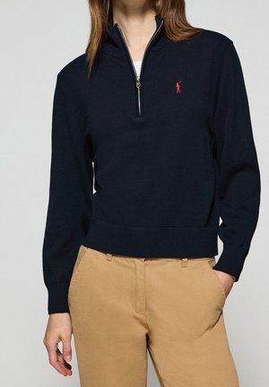 Personne portant un pull bleu marine à fermeture éclair quart de zip avec un logo rouge brodé et un pantalon beige, mains détendues sur les côtés.