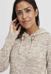 Beige strikket hettegenser med teksturert mønster, snorjustering med sirkulære brune detaljer, og ribbestrikkede mansjetter.