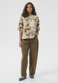 Frau mit lockigem Haar trägt ein beige-blaues Batikshirt, braune Hose und blaue Ballerinas, steht mit einer Hand in der Tasche vor weißem Hintergrund.