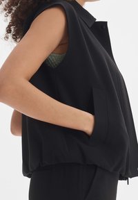 Czarna bezrękawnik z kołnierzykiem, przednimi kieszeniami i marszczonym dołem. Materiał wydaje się gładki z delikatnym połyskiem.