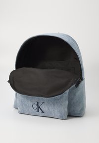 Sac à dos en denim bleu clair avec ouverture zippée. Il dispose d'un intérieur en tissu noir et d'une poche avant avec le logo "CK" en marine.