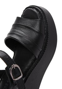 Sandale à plateforme en cuir noir avec une large bride sur les orteils et une bride de cheville ajustable avec boucle argentée, présentée sur fond blanc.