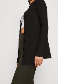 Kvinna klädd i en svart knappad kardigan med utsvängda ärmar, vit kort topp och olivfärgade ribbade byxor mot en neutral bakgrund.