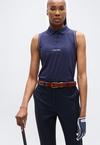 Γυναίκα που φοράει μπλε σκούρο αμάνικο πόλο με λογότυπο "Calvin Klein", σκούρο παντελόνι, καφέ ζώνη και γάντι γκολφ με σχέδιο, κρατώντας ένα μπαστούνι γκολφ.