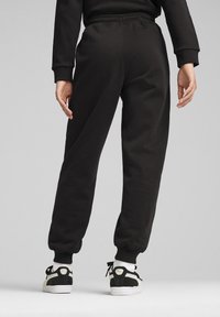 Pantalones de chándal negros con cintura elástica y tobillos ajustados, hechos de una tela suave. Una persona con zapatillas negras y blancas está de pie detrás.