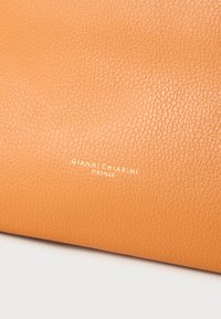 Oranžas ādas tekstūra ar graudainu virsmu. Virsmas centrā atrodas zelta karstumā iespiests logotips "GIANNI CHIARINI FIRENZE".