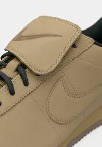 Tenisi bej cu un superior din piele netedă, șireturi negre, logo Nike proeminent pe limba încălțămintei și talpă din cauciuc texturată. Designul tăvălit adaugă structură.