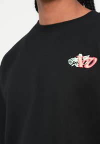 Sudadera negra con cuello redondo, puños de canalé y un gráfico en el lado izquierdo del pecho con letras de gran tamaño en tonos rosa y verde con acentos florales.