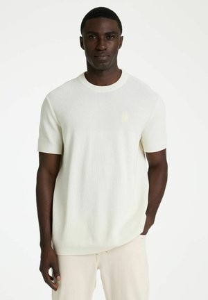 Fekete férfi áll, egyik kezét a zsebében tartja, rövid ujjú fehér kötött pólót és világos színű nadrágot visel, egyszínű háttér előtt.