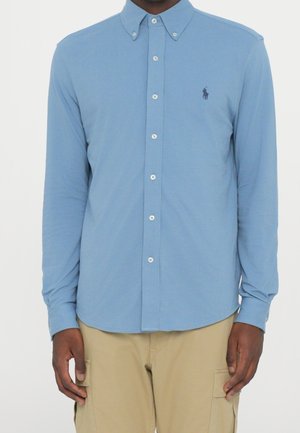 Homme portant une chemise bleu clair boutonnée avec un petit logo brodé sur la poitrine et un pantalon cargo beige.