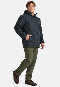 Hombre vistiendo una chaqueta negra con capucha, pantalones oliva y botas de senderismo marrones, de pie con una mano en el bolsillo de la chaqueta.