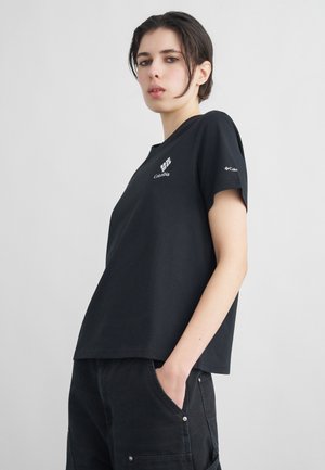 Mladý človek s krátkymi tmavými vlasmi, oblečený v čiernom tričku Columbia a čiernych nohaviciach, stojaci s jednou rukou v kapsičke pred jednoliatym pozadím.