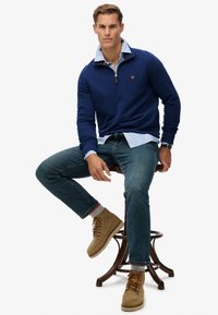 Suéter de cuello con cremallera en azul marino sobre una camisa de rayas en azul claro, combinado con jeans de mezclilla oscura y botas de cordones en marrón claro, sentado en un taburete de madera.