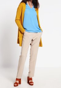 Femme portant un cardigan jaune moutarde, un chemisier bleu, un pantalon beige et des sandales à talons blocs couleur rouille, debout les mains dans les poches.