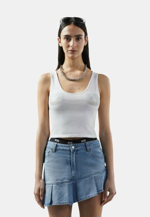 Donna che indossa un top corto bianco senza maniche con decorazioni di perle, gonna di jeans asimmetrica blu, collana a catena argentata e occhiali da sole sulla testa.