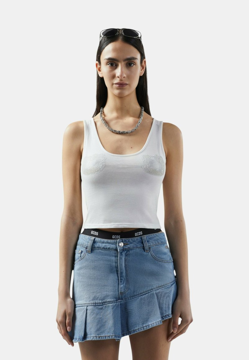 Vrouw die een witte mouwloze cropped top met parelversieringen draagt, een asymmetrische blauwe denimrok, een zilveren ketting en een zonnebril op haar hoofd.