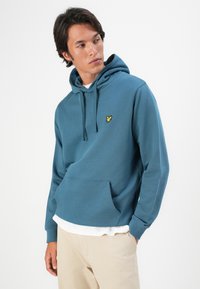 Uomo che indossa un felpa turchese con logo di un uccello giallo, una maglietta bianca sotto e pantaloni beige, che guarda di lato su uno sfondo bianco.