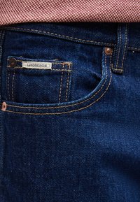 Κοντινό πλάνο στην τσέπη στενών σκούρων μπλε τζιν με καφέ ραφές και μια μικρή λευκή ετικέτα που γράφει "LINDBERGH" κάτω από ροζ υφασμάτινο πουκάμισο.