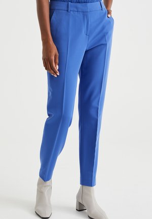 Osoba ubrana w jasnoniebieskie dopasowane spodnie i jasnoszare botki za kostkę, stojąca z jedną ręką w kieszeni na białym tle.