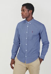 Blauw-witte gestreepte overhemd met lange mouwen, kraag en een klein rood logo op de borst, gedragen met beige broek.
