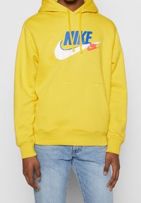 Person som bär en gul hoodie med blå och röda Nike-logotyper, kombinerad med ljusblå jeans, stående mot en enkel bakgrund.