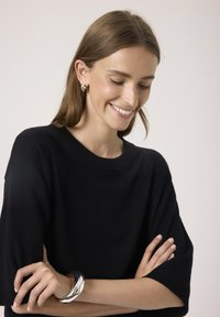 Sort strikket sweater med en afslappet pasform og korte ærmer; model bærer et skinnende sølvmanchetarmbånd og små sølvøreringe.