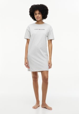 Młoda kobieta stoi boso, ubrana w jasnoszarą sukienkę typu t-shirt z krótkim rękawem, z logo "Tommy Hilfiger" na piersi.